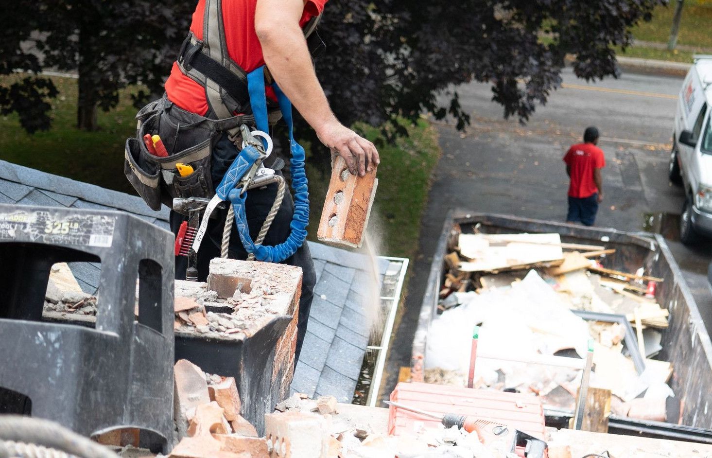 Worker in safety harness removes bricks from roof into dumpster below.